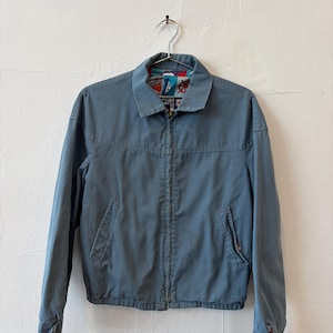 May include: A slate blue zip-up jacket featuring a pointed collar, long sleeves with buttoned cuffs, and two front slash pockets. The interior lining displays a colorful nautical-themed pattern with red, white, and blue maritime signal flags.