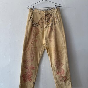 Può includere: Pantaloni in velluto a coste beige con illustrazioni disegnate a mano e scritte rosse. Il design include il disegno di una creatura vicino alla vita, con il testo "PSYCHEDELIC" sulla gamba sinistra e "CUBES" sulla gamba destra.
