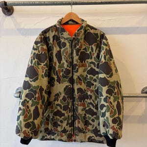 May include: A reversible zip-up jacket featuring a camouflage pattern in shades of brown, green, and black on one side and a solid bright orange color on the other. The jacket has a high collar, long sleeves with black ribbed cuffs, and two front pockets.