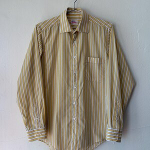 May include: A long-sleeve button-down shirt featuring a vertical striped pattern in mustard yellow and white. The shirt includes a pointed collar, a single chest pocket on the left side, and buttoned cuffs. A label with the text "Jules" is visible.