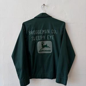 May include: A dark green work jacket featuring a collar and long sleeves. The back displays the text "BRUGGEMAN CO. SLEEPY EYE" in light green lettering above a rectangular patch with a leaping deer graphic and the "JOHN DEERE" logo.