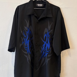 May include: A black short-sleeve button-down shirt featuring a prominent blue and dark grey tribal flame graphic on the front. The shirt has a classic collar, a button-up front closure, and a small brand tag on the lower side seam.