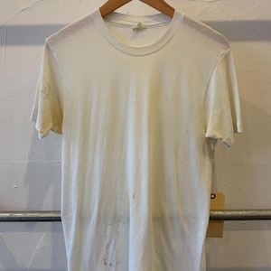 May include: A short-sleeved, off-white crew neck t-shirt featuring a ribbed collar and visible light brown stains scattered across the front and sleeves. The garment is displayed on a wooden hanger and includes a small, blank tan paper tag.