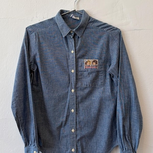May include: A blue chambray long-sleeve button-down shirt with a pointed collar and a single chest pocket. The pocket features a small rectangular patch with an illustration of two dolls and the text Cabbage Patch Kids in red lettering.