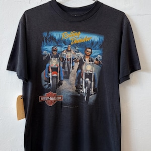 May include: A black short-sleeve t-shirt featuring a graphic print of three motorcyclists riding on a road. The text Rolling Thunder appears in yellow above the riders, and the Harley-Davidson Motor Cycles logo is printed in orange and white below.