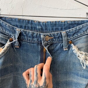 K&ouml;nnte beinhalten: Nahaufnahme einer blauen Jeans mit Knopf- und Rei&szlig;verschluss. Die Jeans hat abgenutzte Details an den Kanten und Taschen. Die Jeans h&auml;ngt an einem Metallb&uuml;gel.