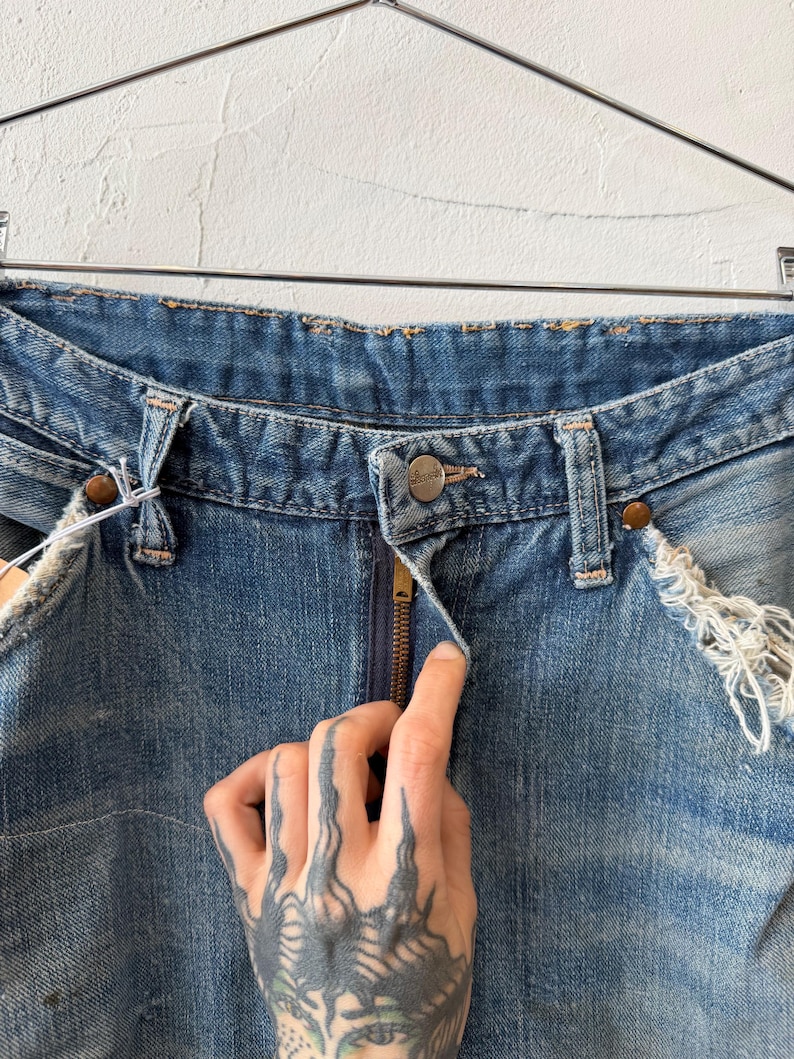 K&ouml;nnte beinhalten: Nahaufnahme einer blauen Jeans mit Knopf- und Rei&szlig;verschluss. Die Jeans hat abgenutzte Details an den Kanten und Taschen. Die Jeans h&auml;ngt an einem Metallb&uuml;gel.