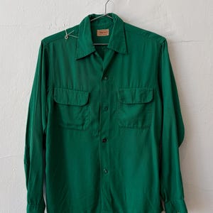May include: A long-sleeve button-down shirt in a solid emerald green color. The shirt features a pointed collar, two chest pockets with buttoned flaps, and a central button placket. A small white tag is attached to the right shoulder area.