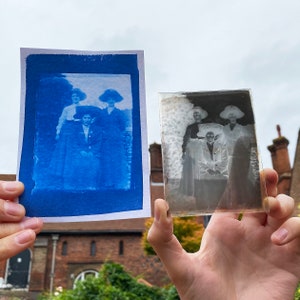 May include: A cyanotype print of a group of four people wearing hats. The print is held in a person's hand. The original glass negative is also held in a hand. The glass negative is a black and white image of the same group of people.