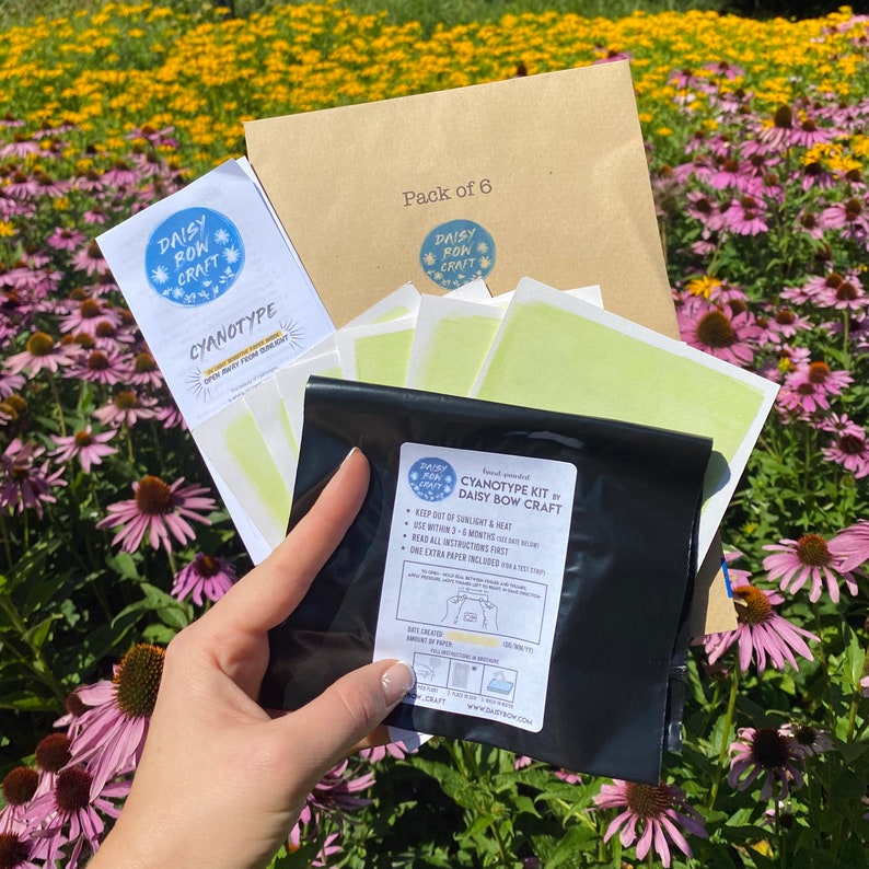 May include: A cyanotype kit by Daisy Bow Craft, packaged in a black plastic bag with a white label. The label includes instructions for use and a list of materials. The kit is displayed on a table with a brown envelope and a pack of six cyanotype paper sheets.