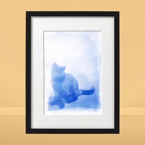 Op de afbeelding: Een ingelijste foto van een kat die zit en omhoog kijkt. De kat is in een zachte blauwe tint, en de achtergrond is lichtblauw.