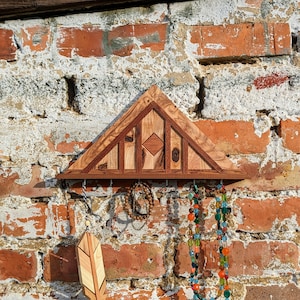 以下が含まれることがあります： 幾何学模様の三角形の木製壁掛けオーガナイザー。ネックレス、ブレスレット、その他のジュエリーを掛けるためのフックが3つ付いています。木製のはねのチャームがフックの1つにぶら下がっています。