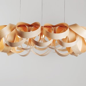 May include: A modern, hanging light fixture with a unique design. The fixture is made of layered, curved wooden strips, creating a flowing, organic shape. The light has a warm, natural wood tone and includes several light bulbs. The fixture is suspended from the ceiling.
