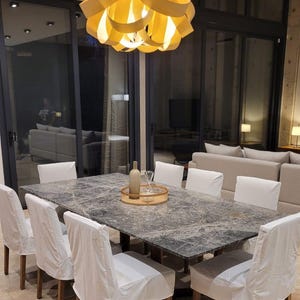 May include: A rectangular dining table with a dark gray marble top and eight chairs featuring white fabric slipcovers. Above the table hangs a large, circular pendant light fixture composed of overlapping yellow ribbons emitting a warm glow.