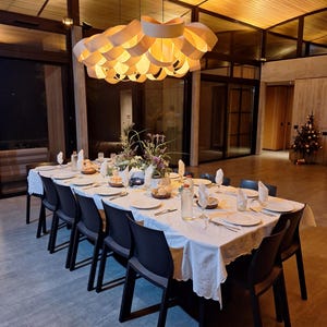 May include: A long dining table set for a meal, covered with a white tablecloth and set with plates, glasses, and silverware. Black chairs are arranged around the table. A large, decorative light fixture hangs above the table.