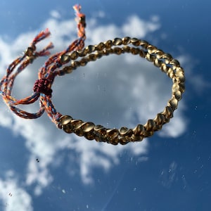 May include: A gold-colored bracelet with a textured design, resting on a reflective surface. The bracelet is connected to a colorful woven cord with a knotted detail. The background shows a sky with clouds.