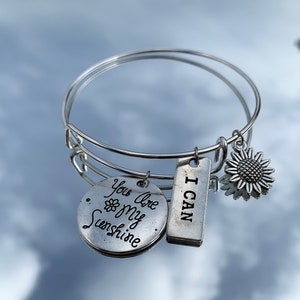 Puede incluir: Pulsera de brazalete plateada con tres colgantes. Un colgante es un disco redondo con las palabras "You Are My Sunshine" y un girasol. Otro colgante es una etiqueta rectangular que dice "I CAN". El tercer colgante es un girasol.