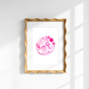 May include: A rectangular wooden frame with a wavy, scalloped edge containing a white print. The print features a circular watercolour illustration in shades of pink, depicting a textured moon-like sphere adorned with small, vibrant pink flowers.