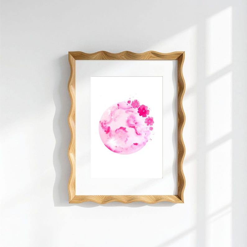 May include: A rectangular wooden frame with a wavy, scalloped edge containing a white print. The print features a circular watercolour illustration in shades of pink, depicting a textured moon-like sphere adorned with small, vibrant pink flowers.