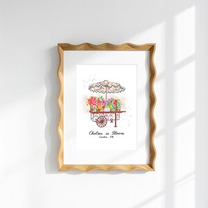 K&ouml;nnte beinhalten: Ein gerahmter Kunstdruck mit einer Aquarellillustration eines Blumenwagens mit einem blumig gemusterten Sonnenschirm. Der Wagen ist mit bunten Blumen gef&uuml;llt und zeigt unterhalb den Text Chelsea in Bloom London, UK in einer schwarzen Serifenschrift.
