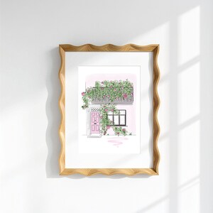May include: A framed art print featuring a line drawing of a building facade with a pink door and a balcony covered in climbing pink roses and green leaves. The frame has a wavy, light brown wooden edge and a white mat border around the print.