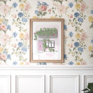 May include: A framed art print featuring a watercolor illustration of a building facade with a pink door, a black balcony, and climbing green vines with pink flowers. The artwork is displayed against a floral wallpaper background with blue, yellow, and pink blossoms.