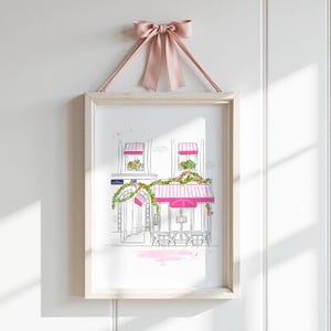 Op de afbeelding: Een ingelijste kunstdruk met een lijntekening van een Parijs caf&eacute; met roze gestreepte luifels, groene klimplanten en zitplaatsen buiten. De print is opgehangen aan een oudroze satijnen strik aan de bovenkant van de houten lijst.