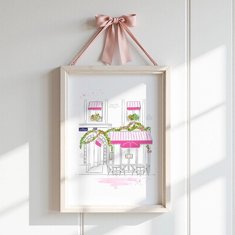 Op de afbeelding: Een ingelijste kunstdruk met een lijntekening van een Parijs caf&eacute; met roze gestreepte luifels, groene klimplanten en zitplaatsen buiten. De print is opgehangen aan een oudroze satijnen strik aan de bovenkant van de houten lijst.