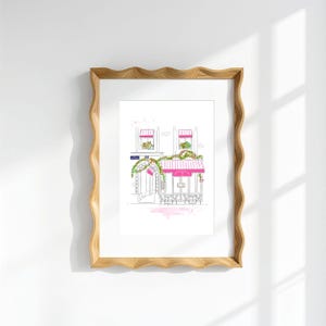 Op de afbeelding: Een ingelijste lijntekening van een caf&eacute;gevel in Parijse stijl met roze gestreepte luifels, groene klimplanten en zitplaatsen buiten. Het kunstwerk zit in een lichtgekleurde houten lijst met golvende randen tegen een effen witte muur.