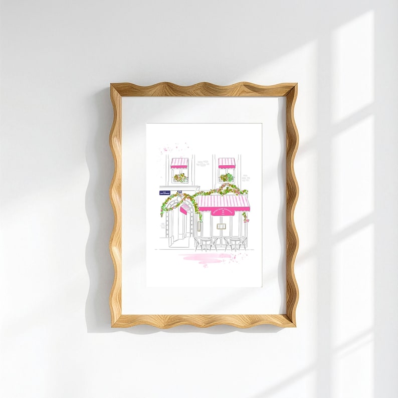 Op de afbeelding: Een ingelijste lijntekening van een caf&eacute;gevel in Parijse stijl met roze gestreepte luifels, groene klimplanten en zitplaatsen buiten. Het kunstwerk zit in een lichtgekleurde houten lijst met golvende randen tegen een effen witte muur.