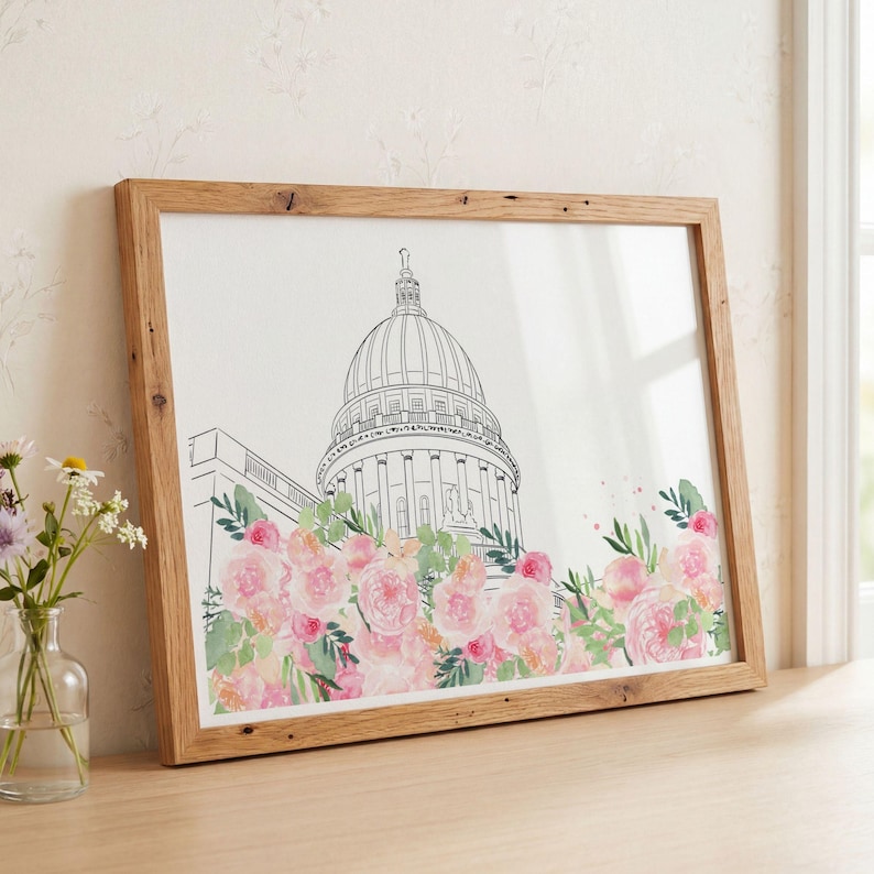 May include: A framed art print featuring a black line drawing of a domed capitol building. The base of the structure is surrounded by a watercolor illustration of pink and peach roses with green leaves, set within a light brown wooden frame.