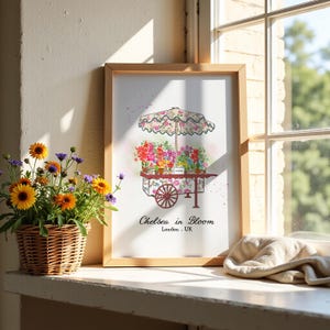 K&ouml;nnte beinhalten: Ein gerahmter Kunstdruck mit einer Aquarellillustration eines Blumenwagens mit gemustertem Sonnenschirm und verschiedenen Topfblumen. Der Text lautet Chelsea in Bloom London, UK. Der Druck befindet sich in einem hellen Holzrahmen.