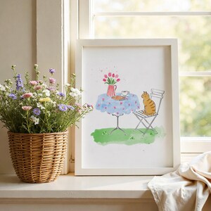 May include: A framed watercolor print featuring a ginger cat sitting on a white metal chair next to a round table with a blue floral tablecloth, a red and white striped pitcher of pink tulips, and a book, all set on a patch of green grass.