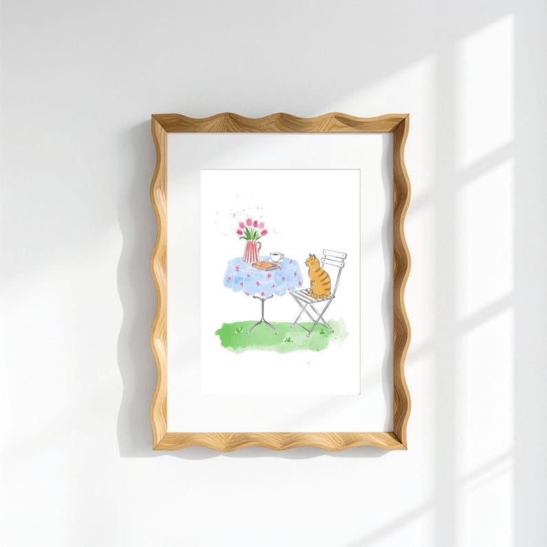 May include: A framed watercolor illustration featuring a ginger tabby cat sitting on a white metal chair next to a round table with a blue floral tablecloth. On the table sits a red and white striped pitcher with pink tulips and a small cup.
