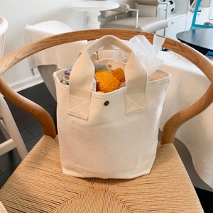 May include: A white canvas tote bag with two thick handles. The bag is filled with items, including a stuffed animal and a few bottles.