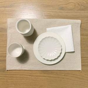 May include: A white ceramic plate with a fluted design sits on a beige linen placemat. Two white ceramic cups are also on the placemat. A white cloth napkin is on the placemat.