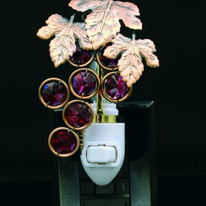 Puede incluir: Una luz nocturna con forma de vid con hojas de cobre y uvas de cristal morado. La luz nocturna se conecta a una toma de corriente estándar.
