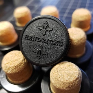 May include: A collection of bottle stoppers with cork tops and black bases. One stopper is in focus, featuring the word "HENDRICK'S" and a fleur-de-lis design. The stoppers are arranged on a dark surface.