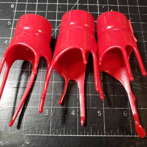 May include: Three bright red plastic objects with a cylindrical top and a dripping, elongated bottom. The objects are arranged vertically, with the dripping sections pointing downwards. The background is a dark surface with grid lines.