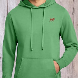 May include: A bright green long-sleeve hooded sweatshirt featuring a small, embroidered brown dog graphic on the left chest. The garment includes a front kangaroo pocket, a drawstring hood with matching green cords, and ribbed cuffs and hem.
