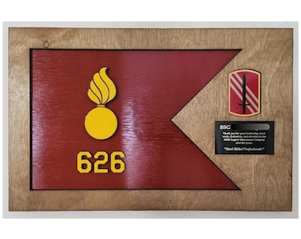 May include: A framed wooden plaque featuring a red flag design with a yellow flame and circle emblem, and the number 626 in yellow. A smaller emblem and a black plaque with text are also present. The frame is light brown.