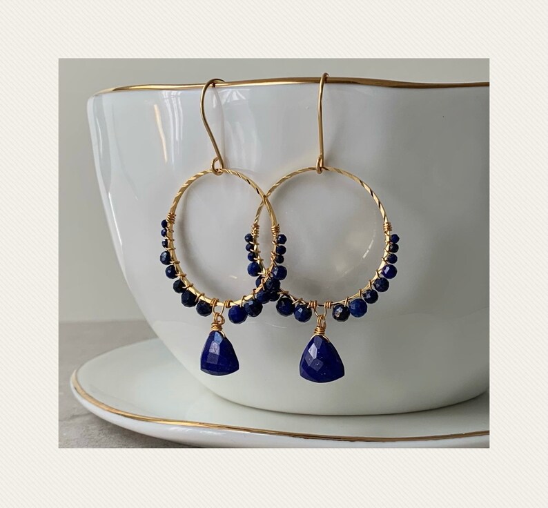 May include: Gold hoop earrings with a dangling lapis lazuli gemstone. The hoops are adorned with small lapis lazuli beads.
