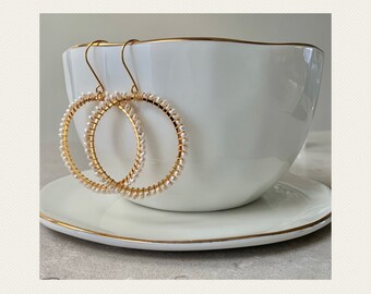 May include: A pair of gold hoop earrings with white pearl accents. The earrings are displayed on a white saucer.
