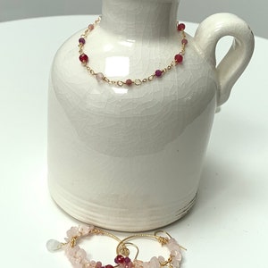 Könnte beinhalten: Ein Set aus goldfarbenen Schmuck mit rosafarbenen und roten Edelsteinperlen. Die Halskette liegt auf einer weißen Keramikvase und die Ohrringe liegen auf einer weißen Oberfläche.