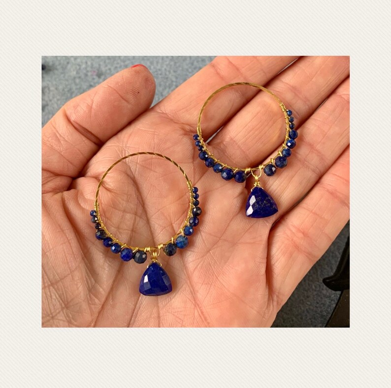 May include: Gold hoop earrings with a dangling lapis lazuli gemstone. The hoops are adorned with small blue lapis lazuli beads.