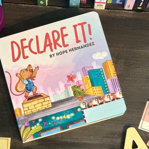 May include: A children's book titled "Declare It!" by Hope Hernandez. The cover features a cartoon mouse standing on a rooftop with a city skyline in the background. The mouse is wearing a blue shirt and red pants.