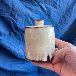 May include: A small, off-white ceramic jar with a brown glaze. The jar has a rounded top and a slightly flared base. The jar is being held in a hand.