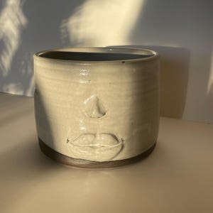 May include: A cylindrical ceramic mug with a face design. The off-white mug features a nose and lips, with a dark brown base and interior. The mug is approximately 4 inches tall and 3 inches in diameter.