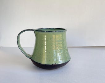 May include: A handmade ceramic mug with a light green exterior and a dark brown base. The mug has a curved handle and a dark blue rim. The mug is designed for hot beverages and is suitable for home or office use.