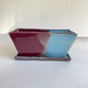 May include: A rectangular ceramic planter with a divided color scheme. One half is a deep burgundy, the other is a light blue. The planter sits on a matching rectangular base. The planter is suitable for small plants or succulents.
